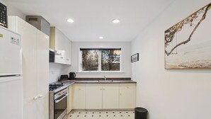 A compact kitchen with white cabinets, a gas stove, a microwave, and a window with a view of trees. - Wanaka Central Holiday Escape (Wanaka)