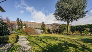 A well-maintained garden with a variety of plants and a large tree, a gravel path, and a view of distant mountains. - Wanaka Central Holiday Escape (Wanaka)