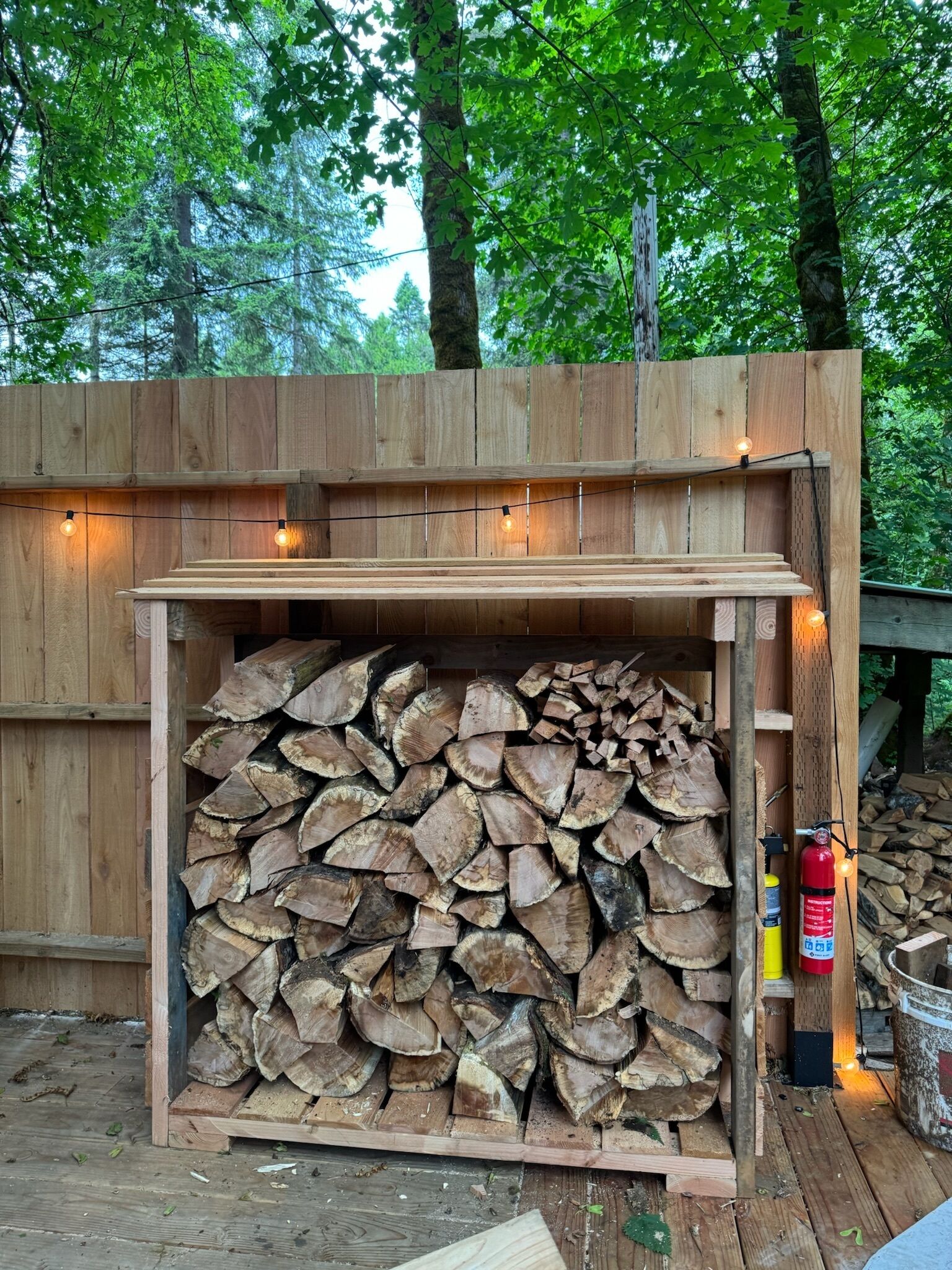 A wooden outdoor fireplace with a stack of firewood, a fire extinguisher, and string lights.