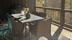 Ein überdachter Sitzbereich im Freien mit Tisch, Stühlen, einem Grill und Blick auf Herbstlaub.