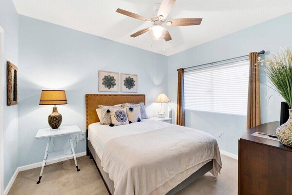 Un dormitorio con cama, ventilador de techo, ventana con persianas, mesita de noche, lámpara y un jarrón decorativo.