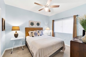 Quarto com cama, ventoinha no teto, janela com persianas, mesa de cabeceira, abajur e um vaso decorativo.