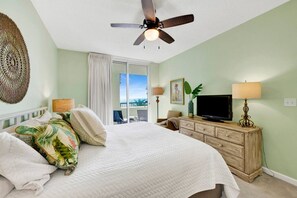 A bedroom with a bed, a television, a dresser, a ceiling fan, and a view of the outdoors.