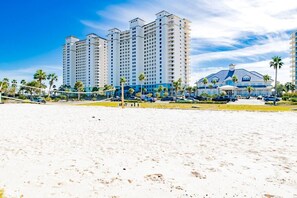 Een strand met een volleybalnet, palmbomen en een hotel met meerdere verdiepingen en balkons.