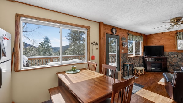 A cozy cabin interior with a wooden dining table, chairs, a stone fireplace, a television, and a large window offering a view of snow-covered trees.