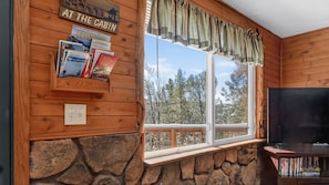 A cabin interior with a stone wall, wooden paneling, a window with a view of trees, a bookshelf, and a sign reading "AT THE CABIN".
