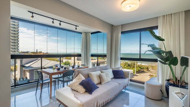 A modern living room with a sofa, a dining table, and chairs, large windows with a view of the sea, and a potted plant. - Fully Equipped and Modern Apartments 100m From the Beach - Rpos (Picarras)