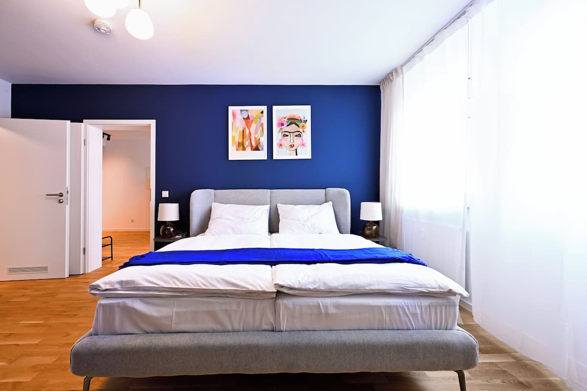 A bedroom with a bed, two bedside tables, a nightstand, a blue wall, and two framed artworks.