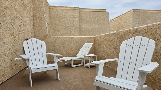 A set of white Adirondack chairs with attached side tables, positioned on a sandy surface against a textured wall.
