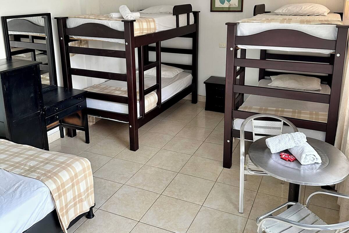 A dormitory room with three bunk beds, a desk, a chair, a small table, and a ceiling fan.