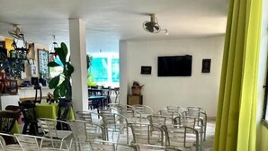 A spacious room with a tiled floor, clear plastic chairs arranged in rows, a large potted plant, and a flat-screen TV mounted on the wall. - HOTEL AVENIDA 15 (Melgar)