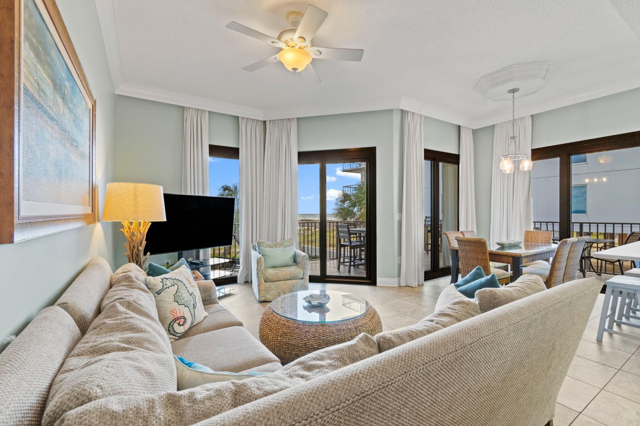 A spacious living room with a sofa, armchair, and a coffee table. There is a television, a ceiling fan, and a balcony with a dining area.