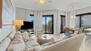 Amplia sala de estar con sofá, sillón y una mesa de centro. Hay un televisor, un ventilador de techo y un balcón con zona de comedor.