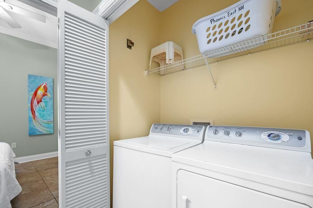 A laundry area with a white washer and dryer, a shelf with a basket labeled "Phoenix East 101," and a painting of a fish on the wall.