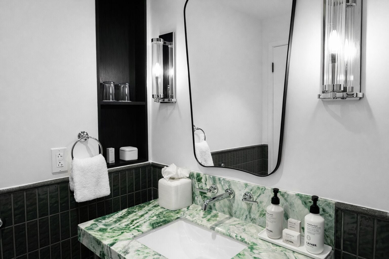 A modern bathroom with a white sink, green marble countertop, and a large mirror. There are toiletries on the counter and a towel hanging on the wall.