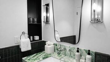 A modern bathroom with a white sink, green marble countertop, and a large mirror. There are toiletries on the counter and a towel hanging on the wall.