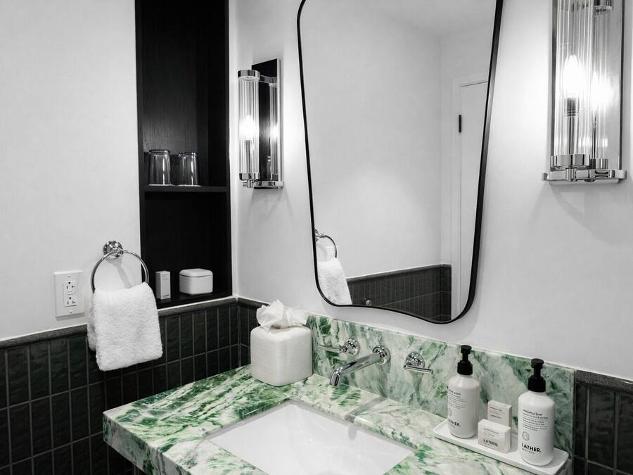 A modern bathroom with a white sink, green marble countertop, and a large mirror. There are toiletries on the counter and a towel hanging on the wall.