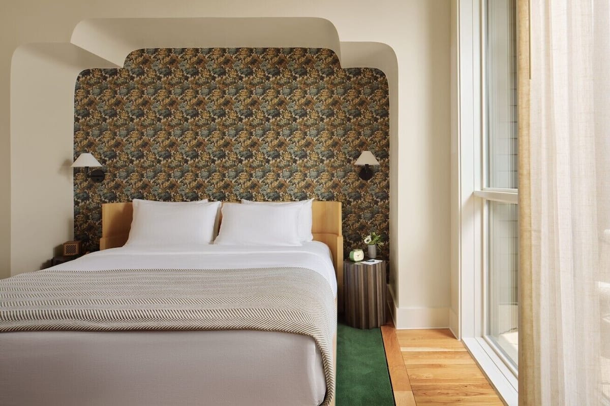Une chambre d’hôtel avec un grand lit, une tête de lit à motifs, deux lampes de chevet, une petite table d’appoint et une fenêtre avec des rideaux.