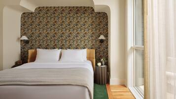 Quarto de hotel com uma cama grande, cabeceira estampada, duas luminárias de cabeceira, mesa de apoio pequena e uma janela com cortinas.