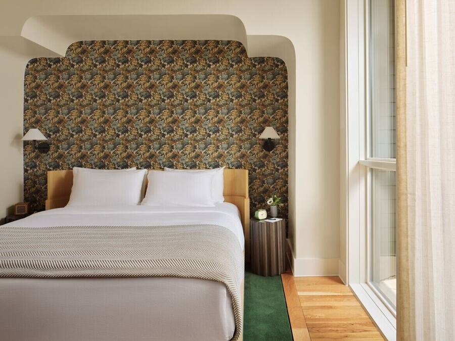 Quarto de hotel com uma cama grande, cabeceira estampada, duas luminárias de cabeceira, mesa de apoio pequena e uma janela com cortinas.