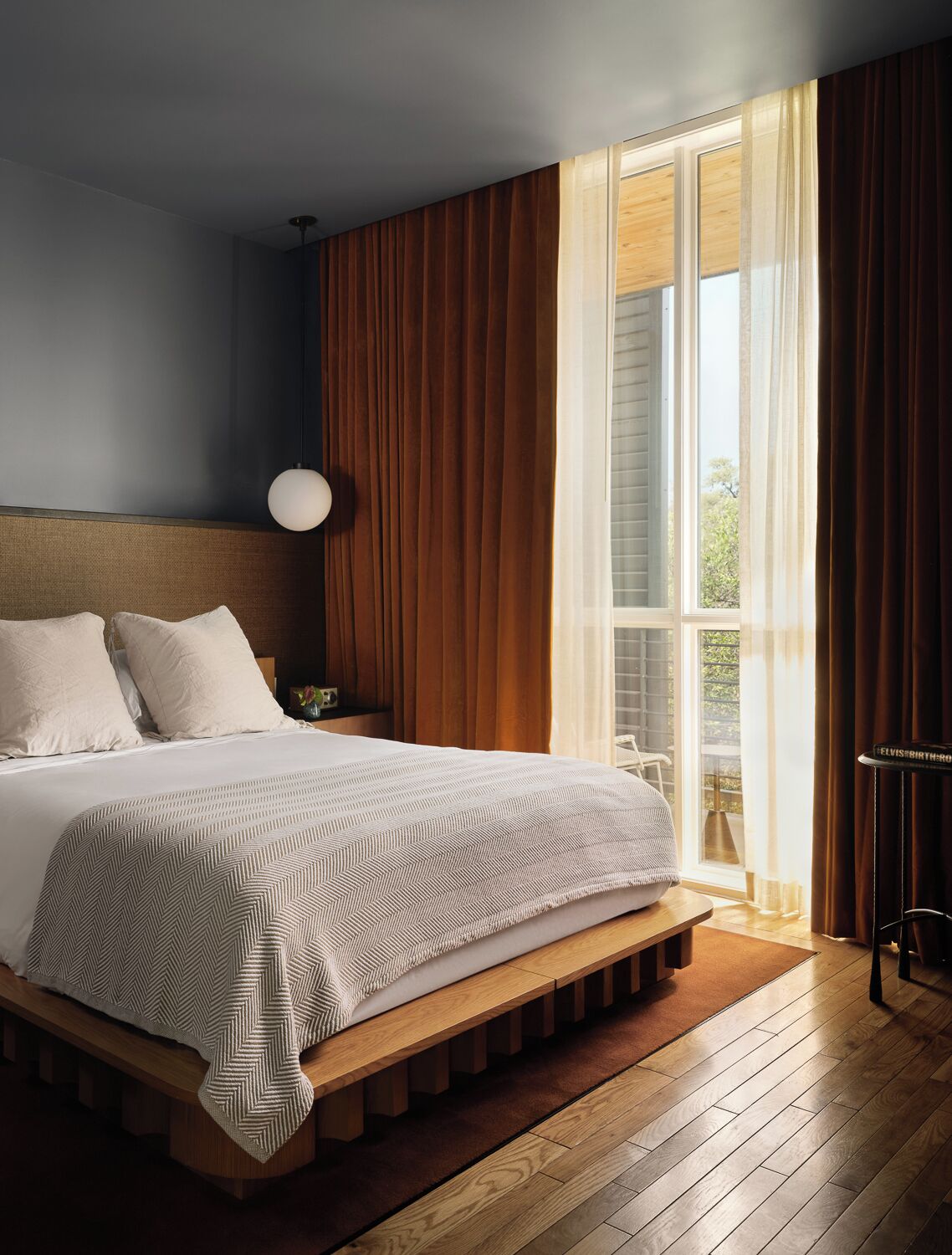 A modern bedroom with a large bed, a wooden headboard, a bedside lamp, and a view of the outdoors through a window.