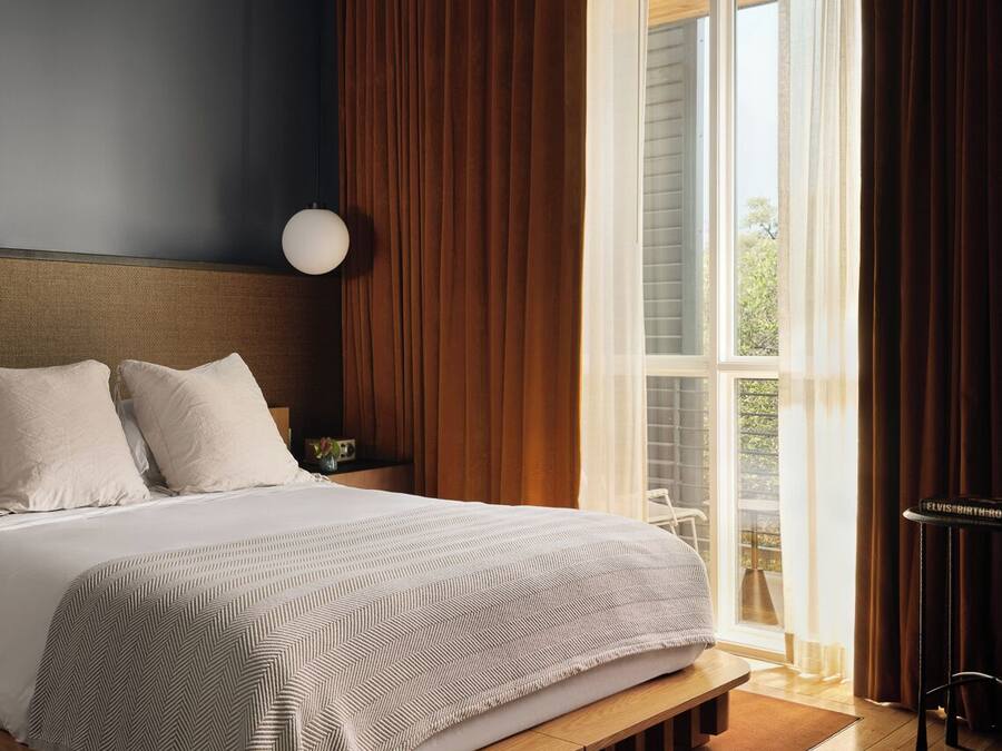 Ein modernes Schlafzimmer mit einem großen Bett, einem Holz-Kopfteil, einer Nachttischlampe und Blick ins Freie durch ein Fenster.