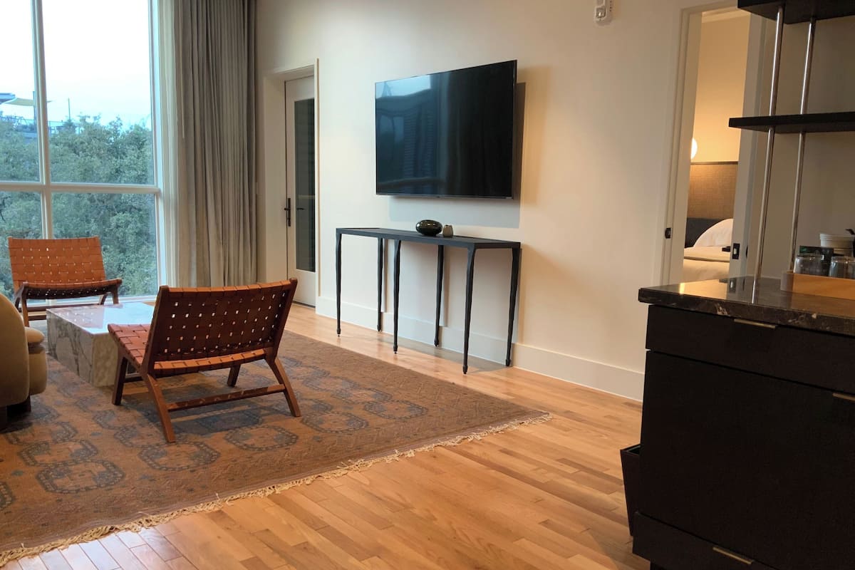Un salon moderne avec une grande fenêtre, un téléviseur à écran plat, une chaise en bois, une petite table et un meuble de couleur sombre.