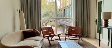 A modern living room with a sofa, two wooden chairs, a marble coffee table, and a view of a building through large windows.