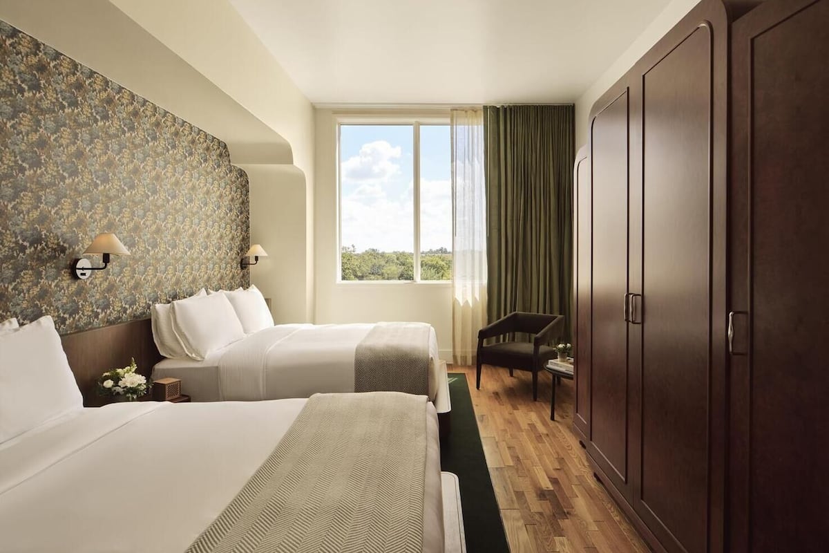 Une chambre d’hôtel avec deux lits, une grande armoire, une fenêtre avec des rideaux et une petite table avec une chaise.
