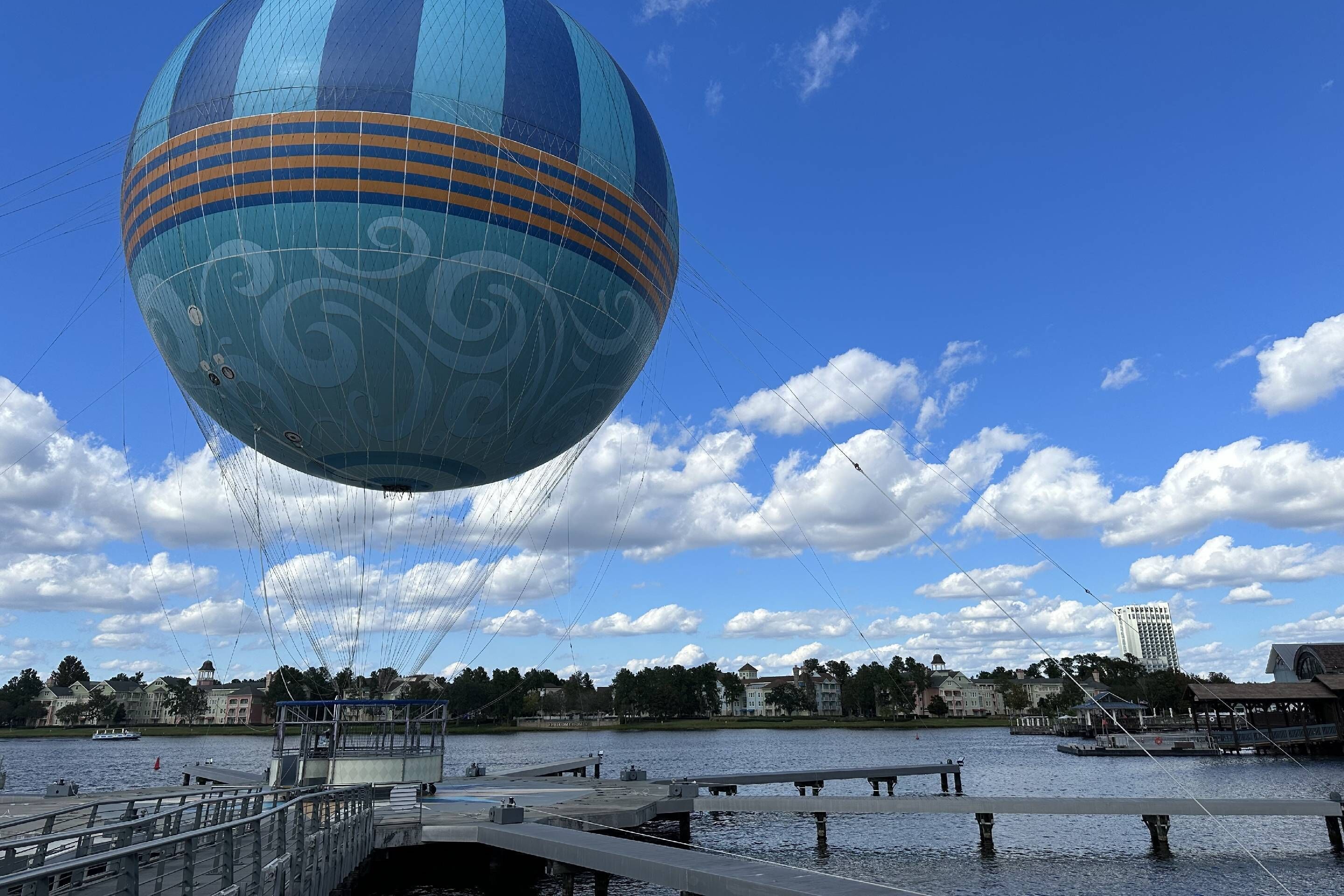 A hot air balloon with a blue and orange design, floating above a waterfront area with buildings and a dock.