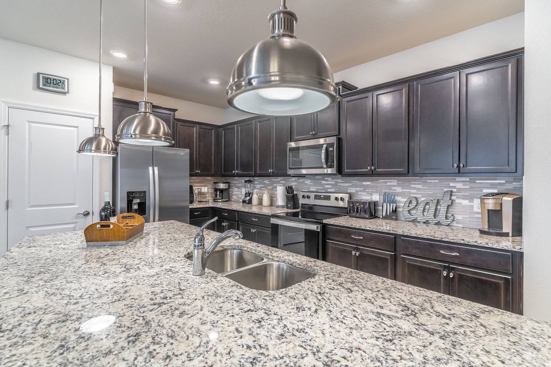 A modern kitchen with dark cabinets, stainless steel appliances, granite countertops, and pendant lighting.