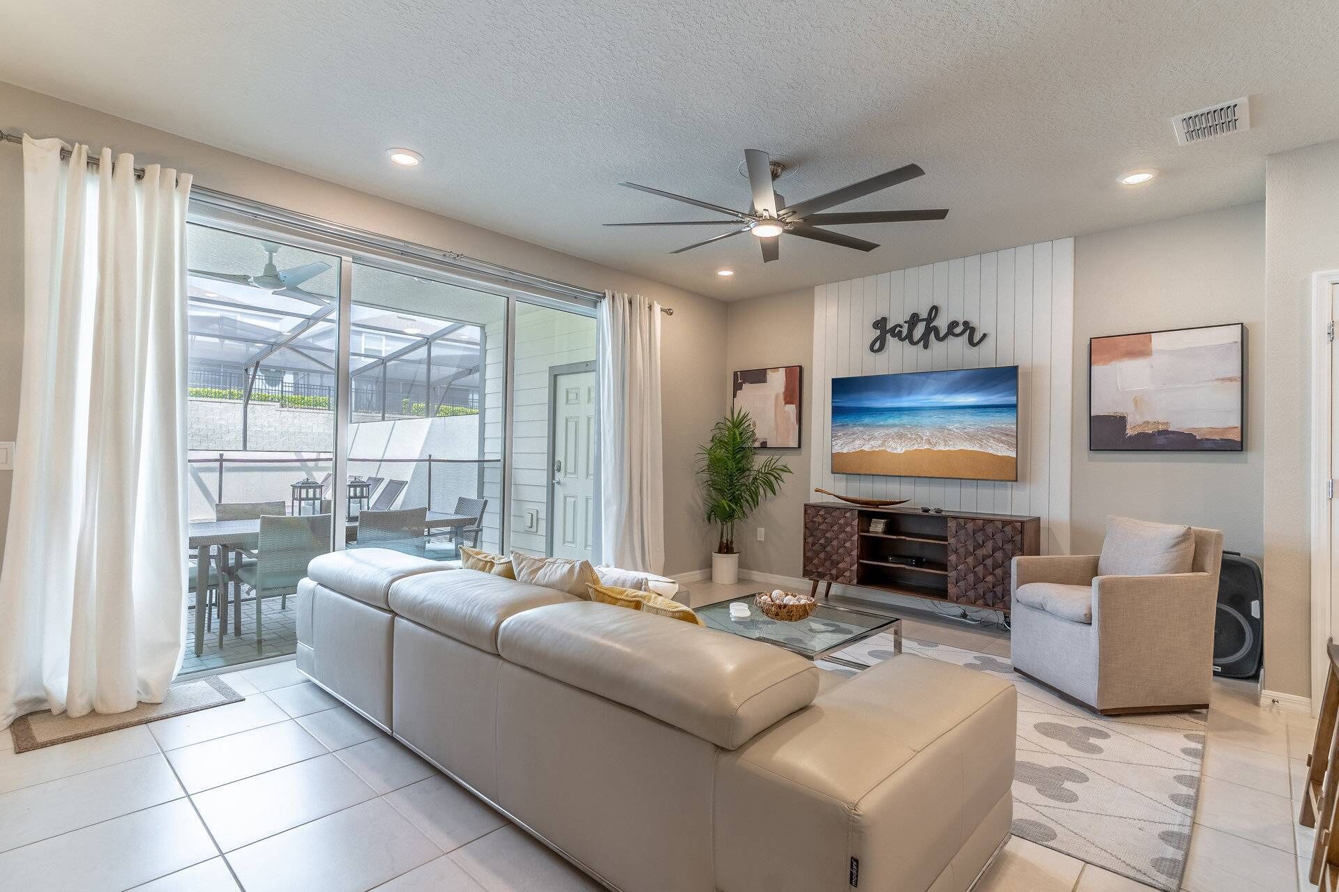 A spacious living room with a large beige sectional sofa, a coffee table, and a television. There is a ceiling fan, a potted plant, and artwork on the walls.