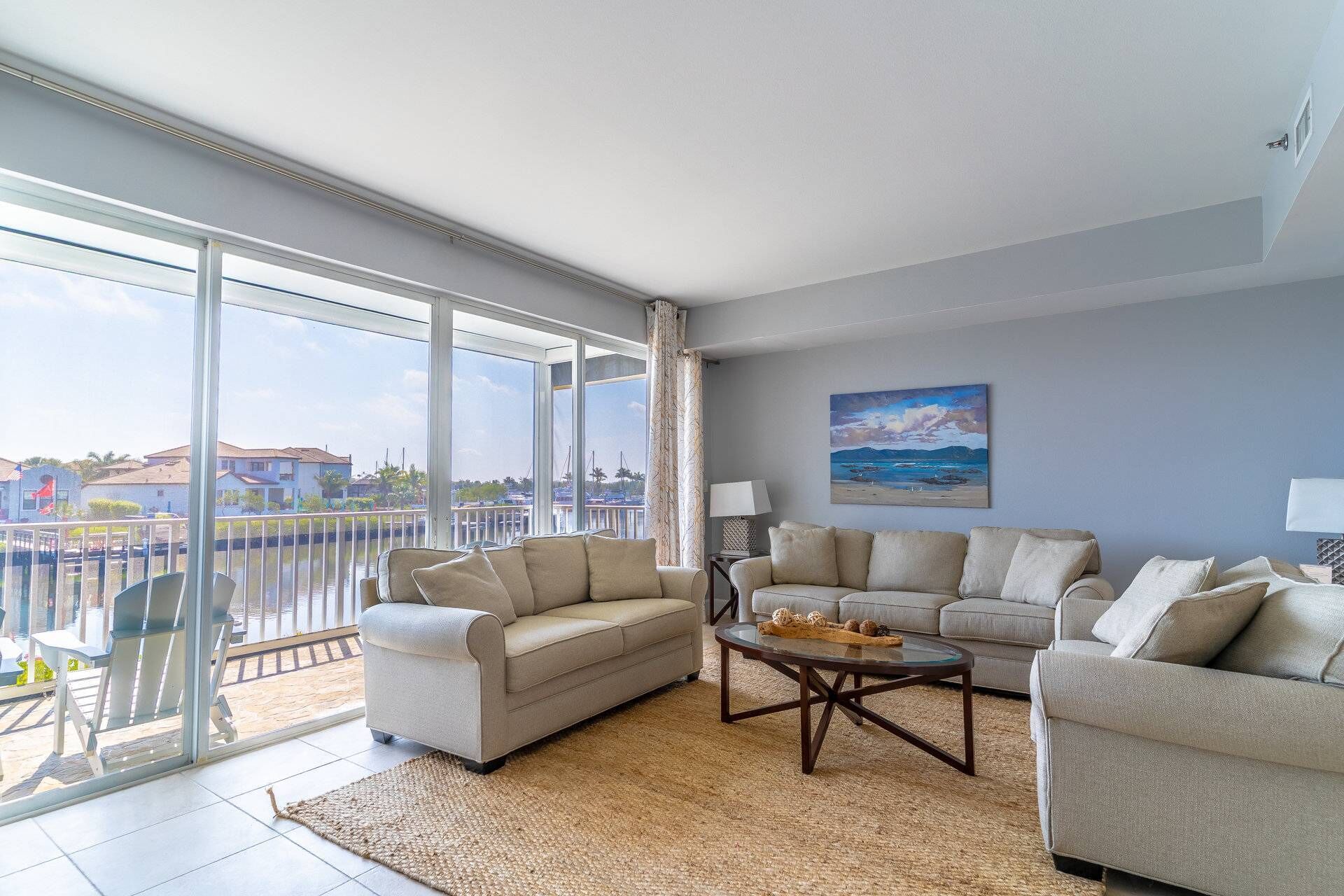 A spacious living room with a large balcony, a sofa set, a coffee table, and a painting of a beach scene.