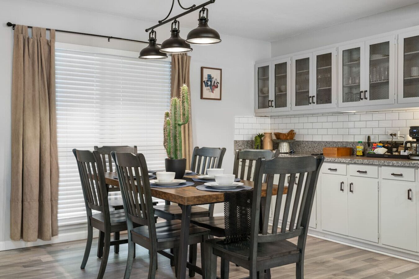 A dining area with a wooden table, chairs, and a potted cactus. There are cabinets with glass doors and a kitchen counter with various items.