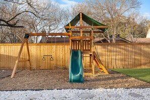 A wooden swing set with a slide, surrounded by a wooden fence and a grassy area.