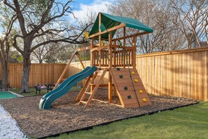 Une structure de jeu en bois, dotée d’un toboggan et d’un mur d’escalade, entourée d’une clôture en bois et d’arbres.