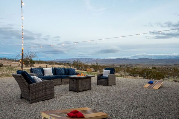 Un espacio al aire libre con mobiliario de mimbre, un hogar de fuego y luces colgantes, con montañas al fondo.