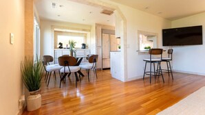 A modern kitchen with white cabinets, a dining area with wooden chairs, and a TV mounted on the wall.