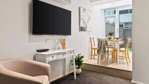 A modern living room with a flat-screen TV, a white console table, a beige armchair, and a dining area with wooden chairs and a table.