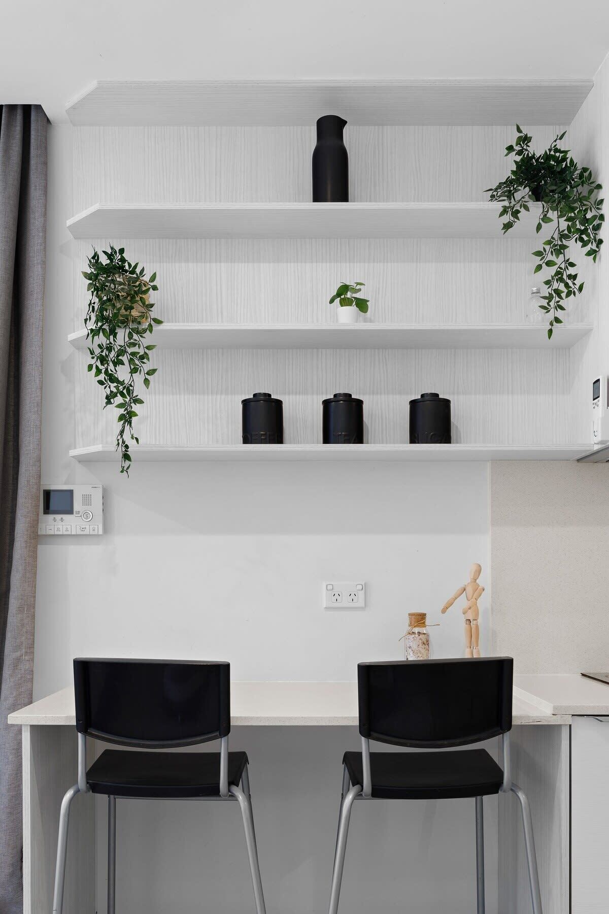 A modern kitchen with white cabinetry, black chairs, and a built-in shelf unit with plants and kitchen appliances.