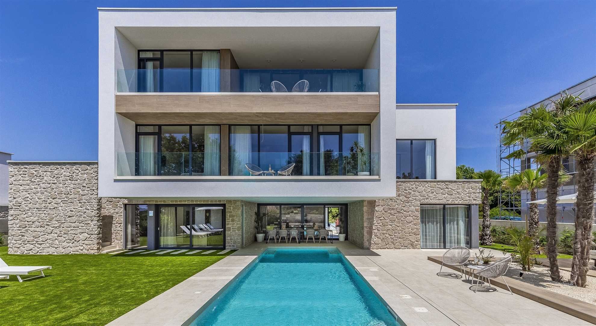 Une maison moderne à deux étages, dotée d’une piscine, de grandes fenêtres et d’une façade en pierre.