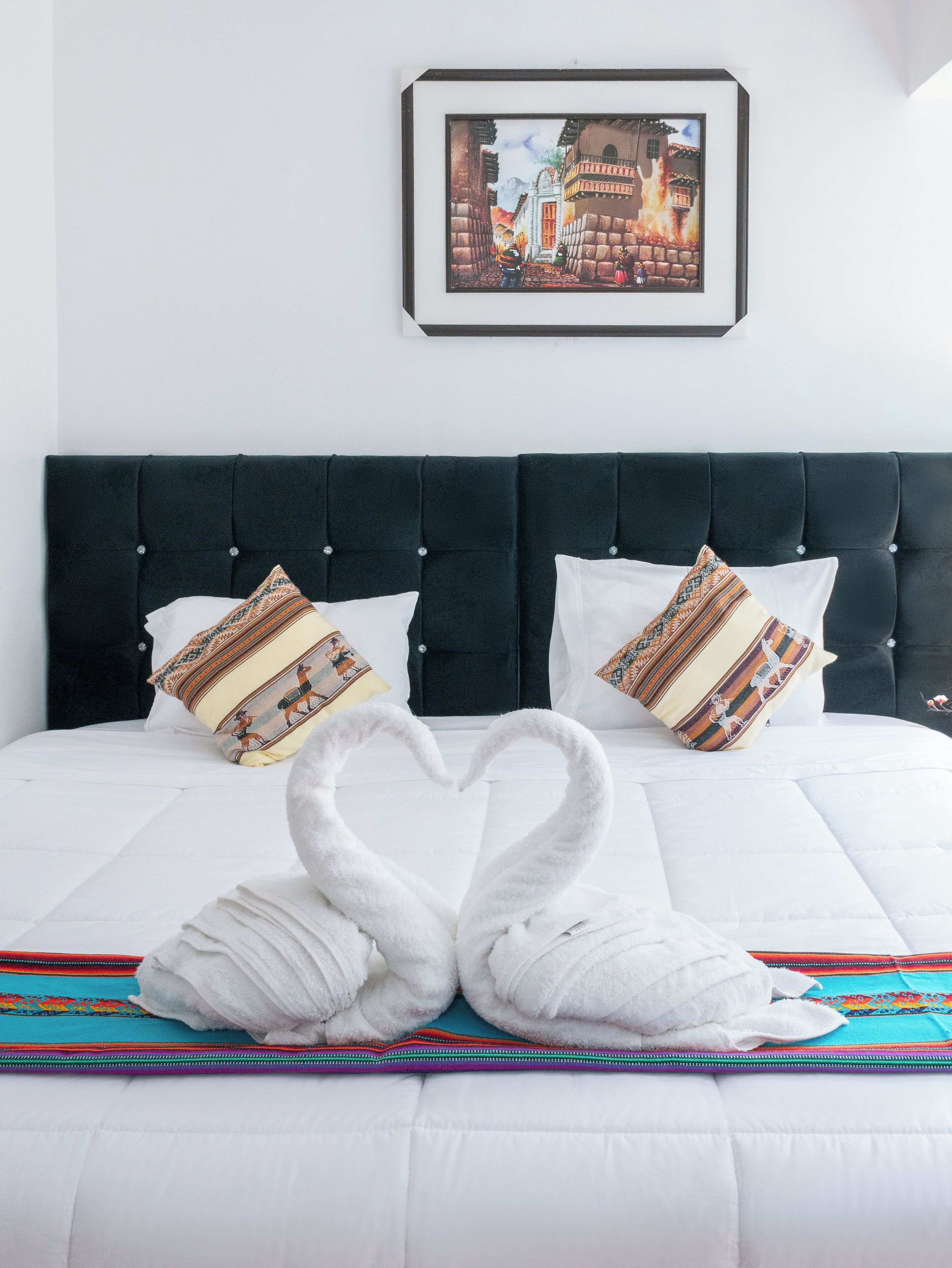 A neatly made bed with a heart-shaped towel arrangement, a framed picture on the wall, and a colorful throw blanket.