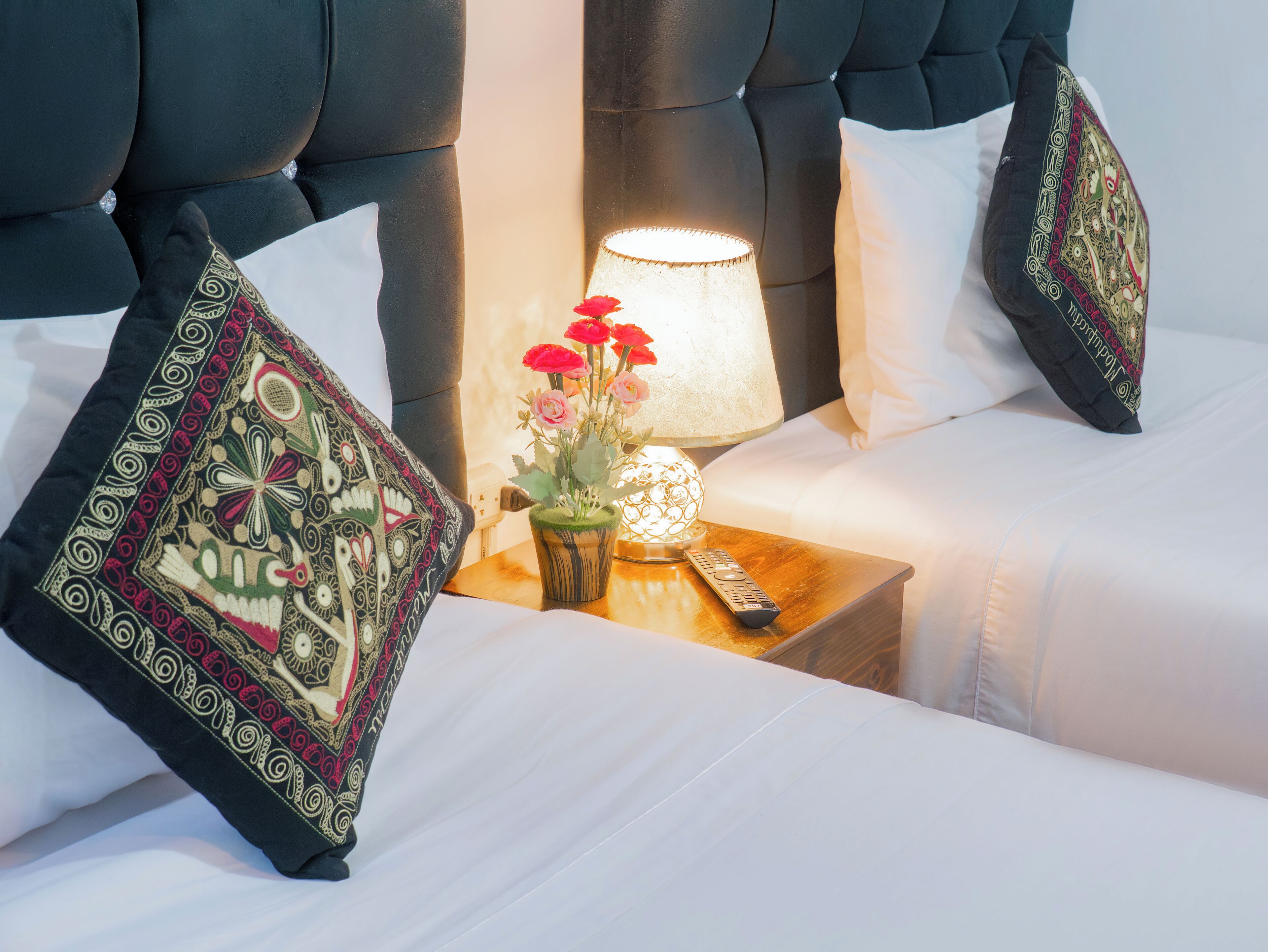 A hotel room with a bed, pillows, a nightstand, a lamp, and a vase with flowers.