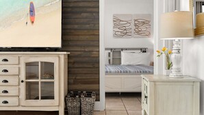 A bedroom with a bed, a bedside table with a lamp, a TV on a wooden cabinet, and a beach scene on the wall. - Beachfront 2BR! · Sunbird 3rd Floor! Free Beach Chairs & Umbrella Service! (Panama City Beach)