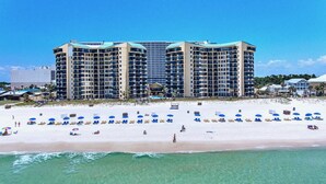 A beachfront hotel with multiple high-rise buildings, a sandy beach with numerous umbrellas and sun loungers, and a clear view of the ocean. - Beachfront 2BR! · Sunbird 3rd Floor! Free Beach Chairs & Umbrella Service! (Panama City Beach)