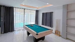 A pool table in a room with a large window, curtains, and a shelf unit. - Anjia Serene Pool Villa (Pattaya)