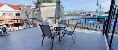 Un balcón con mesa y sillas, una piscina al fondo y un edificio de techo rojo.