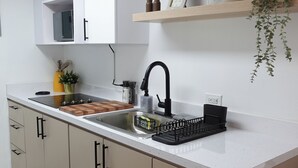 Une cuisine moderne avec un plan de travail blanc, un robinet noir et une planche à découper en bois. On aperçoit une petite plante sur le plan de travail et une étagère au-dessus de l’évier, ornée d’objets décoratifs.