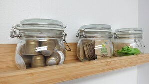 Trois bocaux en verre, posés sur une étagère en bois, contiennent divers objets, comme des pièces de monnaie, du papier et des feuilles vertes.
