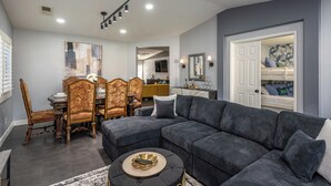 A modern living room with a sectional sofa, a dining table with chairs, and a kitchen area in the background. - Welcome to Villa Tempo - Pool, Spa, Game Room, BBQ, Fast Wifi, Sleeps 12+ (Coachella)